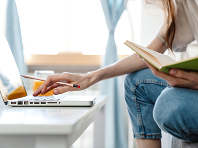 Student working at a laptop, with notebook in hand. Link to Develop your Essay Writing Skills – School of Health and Life Sciences Students.