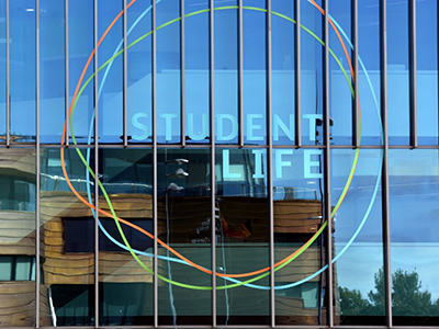 Student Life Building, in sunshine, with the Curve reflected in the window.. Link to Student Life Great Minds Festival Spring 2025.