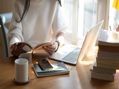 A person reading a book. There is a laptop, a notebook and a pile of books on a desk in front of them. Link to Turn Up and Discover: Postgraduate Study Skills - Thursday 3 April.